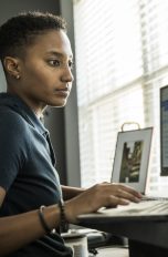 Eine Person sitzt an einem Schreibtisch, auf dem ein Laptop im Vordergrund und ein Monitor dahinter stehen.