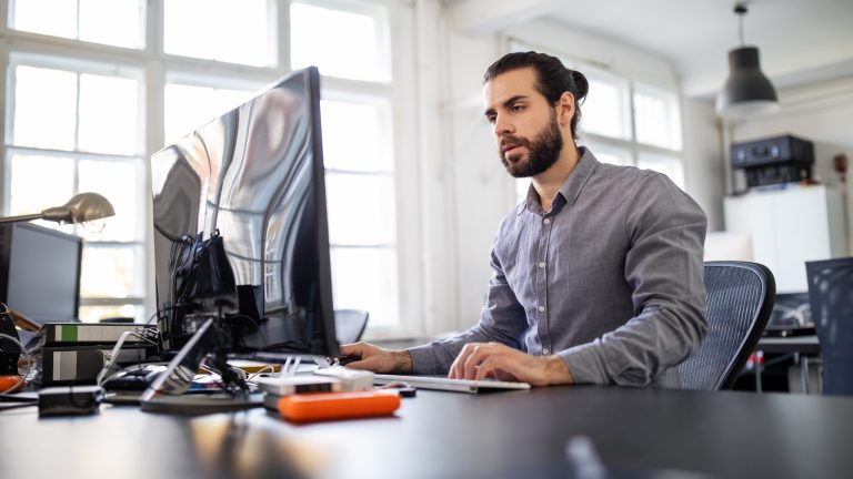 Eine Person sitzt vor einem Computer, Neben dem Bildschirm liegt eine externe Festplatte mit orangefarbiger Schutzhülle. Eine Person sitzt vor einem Computer, Neben dem Bildschirm liegt eine externe Festplatte mit orangefarbiger Schutzhülle.