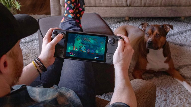 Eine Person sitzt auf einem Sofa und spielt auf einem Valve Steam Deck. Ein auf einem Teppich liegender Hund schaut dabei zu.