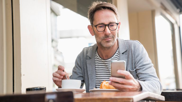 Google Alerts einrichten, verwalten, löschen – ein Leitfaden Eine Person sitzt bei Kaffee und Kuchen draußen an einem Tisch. In der einen Hand hält sie ein Smartphone.