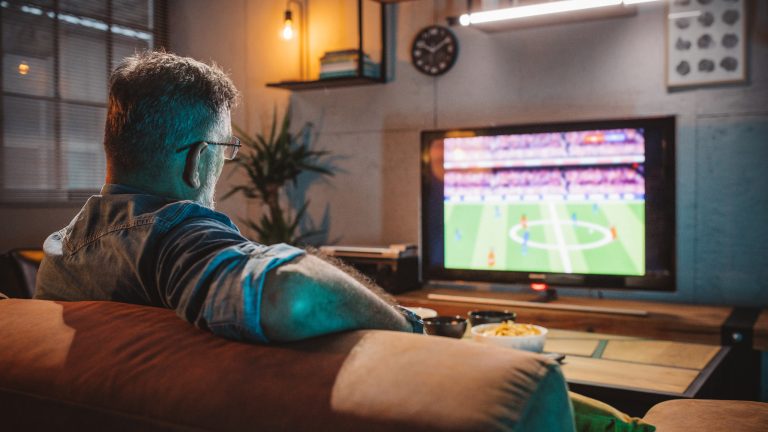 Eine Person sitzt auf einem Sofa und schaut ein Fußballspiel auf einem TV.