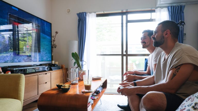 Zwei Personen sitzen auf einem Sofa vor dem Fernseher und halten Controller einer Spielekonsole in der Hand.