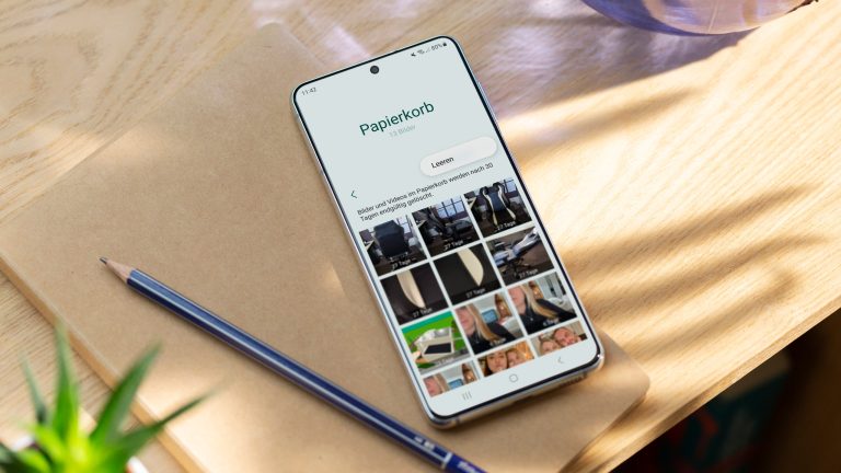 Ein Samsung-Smartphone liegt samt Stift und Notizblock auf einem Holztisch. Im Papierkorb befinden sich mehrere Fotos.
