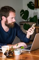 Eine Person sitzt an einem Tisch vor dem Laptop und macht etwas auf ihrem Smartphone.