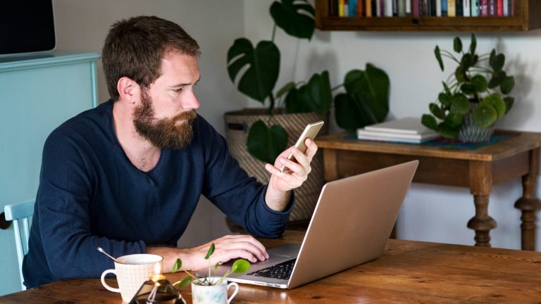 Eine Person sitzt an einem Tisch vor dem Laptop und macht etwas auf ihrem Smartphone.