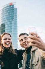 Drei Personen stehen vor hohen Gebäudekomplexen und machen freudestrahlend ein Selfie.