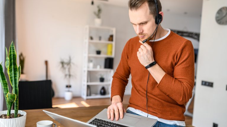 Eine Person sitzt auf einem Tisch und hat einen Laptop auf dem Schoß. Sie trägt ein Headet auf dem Kopf.