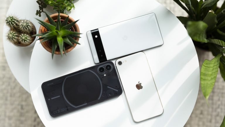 Auf einem weißen Rundtisch liegen ein schwarzes Nothing Phone (1) ein weißes Google Pixel 6a und ein weißes iPhone SE (2022) stilvoll arrangiert.