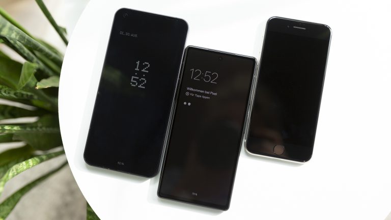 Auf einem weißen Tisch liegen nebeneinander ein Nothing Phone (1), ein Google Pixel 6a und ein iPhone SE (2022). Bei den beiden erstgenannten zeigt ein Always-on-Display Uhrzeit und weitere Informationen an.