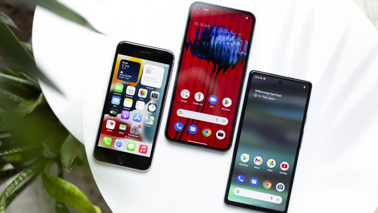 Auf einem weißen Tisch liegen nebeneinander ein Nothing Phone (1), ein Google Pixel 6a und ein iPhone SE (2022). Auf den eingeschalteten Bildschirmen sind die Homescreens zu sehen.