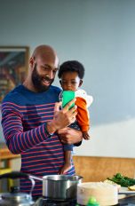 Eine Person mit einem Kind auf dem Arm steht vor einer Kücheninsel. In der freien Hand hält sie ein Smartphone, in das die beiden lächelnd hineinschauen.