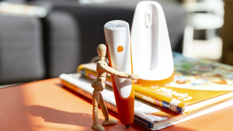 Eine Gliederpuppe hält einen Tiptoi-Stift fest, der neben einem Stapel Tiptoi-Bücher steht. Im Hintergrund ist die dazugehörige Ladestation zu erkennen.