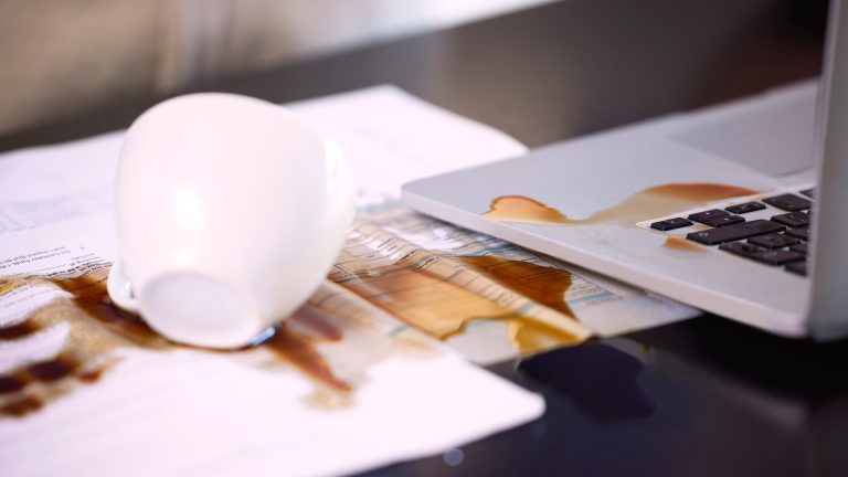 Eine weiße Kaffeetasse liegt samt Inhalt ausgebreitet auf Dokumenten und einer Laptop-Tastatur.
