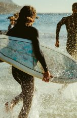 Personen mit Surfbrettern unter dem Arm und Neoprenanzügen laufen ins offene Meer.