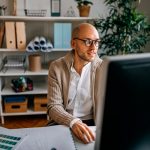 Eine Person sitzt an einem Schreibtisch vor einem Computer und hält eine Maus in der Hand.