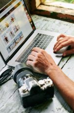 Eine Person sitzt an einem Tisch und schaut auf dem Laptop Bilder an. Daneben liegt eine Spiegelreflexkamera.