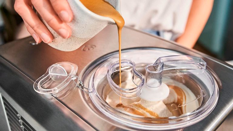 Eine Person gibt eine karamellfarbene Flüssigkeit in eine Eismaschine, während diese die Eismasse rührt. 