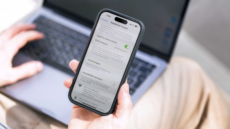 Eine Person hält ein iPhone in der Hand, auf dem gerade ein Hotspot aktiviert wird. Auf dem Schoß der Person ist ein MacBook Pro.