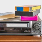 Ein VHS-Player auf einem Schrank. Auf dem Gerät liegen einige VHS-Kassetten.