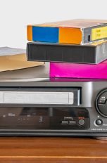 Ein VHS-Player auf einem Schrank. Auf dem Gerät liegen einige VHS-Kassetten.