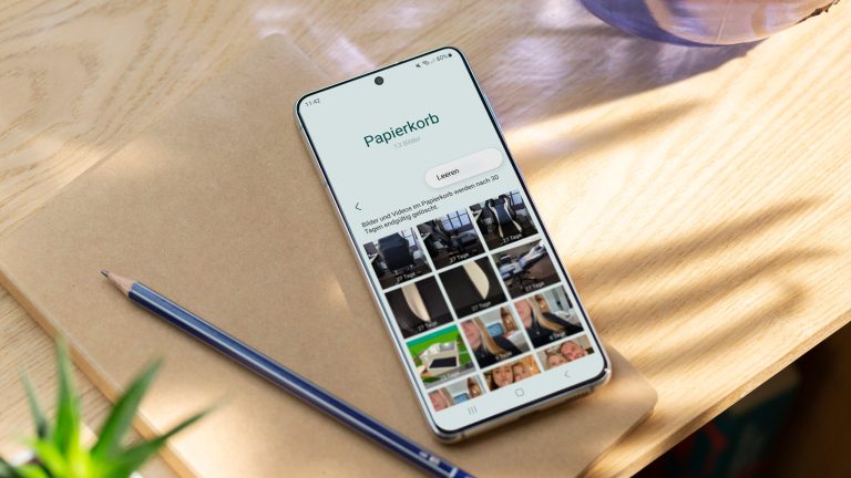Fotos wiederherstellen bei Android, So geht’s Ein Android-Handy mit geöffnetem Papierkorb liegt mit Notizbuch und Stift auf einem Holztisch.