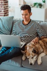 Eine Person sitzt mit einem Hund auf einem Sofa und hat einen aufgeklappten Laptop auf dem Schoß.