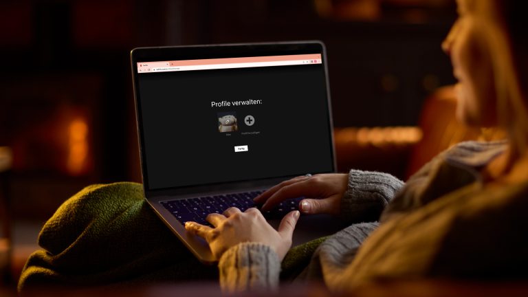 Eine Person sitzt mit einer Decke auf einem Sofa und hat einen Laptop auf dem Schoß. Auf dem Display ist die Profilverwaltung von Netflix zu sehen. Eine Person sitzt mit einer Decke auf einem Sofa und hat einen Laptop auf dem Schoß. Auf dem Display ist die Profilverwaltung von Netflix zu sehen.