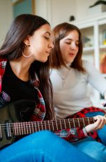 Zwei Personen sitzen auf dem Boden. Eine hält eine E-Gitarre in der Hand, die andere ein Smartphone hoch, auf dem wohl Noten zu sehen sind.