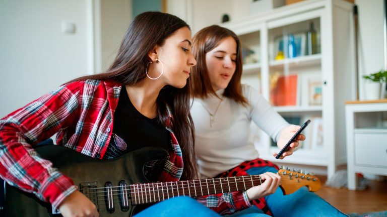 Zwei Personen sitzen auf dem Boden. Eine hält eine E-Gitarre in der Hand, die andere ein Smartphone hoch, auf dem wohl Noten zu sehen sind.