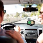 Zwei Personen sitzen in einem Auto. Auf dem Dashboard ist ein Navigationssystem zu sehen.