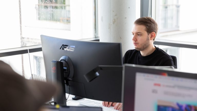 Vor einem Ultrawide-Monitor sitzt eine Person und tippt auf einer Tastatur. Vor einem Ultrawide-Monitor sitzt eine Person und tippt auf einer Tastatur.