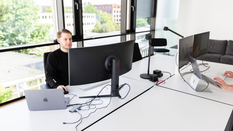 In einem Büro sitzt eine Person vor einem Ultrawide-Monitor. Schräg gegenüber sitzt eine zweite Person vor zwei Monitoren. In einem Büro sitzt eine Person vor einem Ultrawide-Monitor. Schräg gegenüber sitzt eine zweite Person vor zwei Monitoren.
