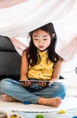 Ein Kind sitzt unter einer Decke, die über ein Sofa gespannt ist und schaut sich etwas auf einem Tablet an.