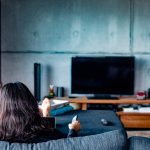 Eine Person sitzt auf einem Sofa und hält eine Fernbedienung Richtung ausgeschaltetem Fernseher.