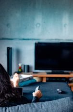 Eine Person sitzt auf einem Sofa und hält eine Fernbedienung Richtung ausgeschaltetem Fernseher.
