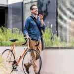 Eine Person führt mit einer Hand ein Fahrrad mit sich, in der anderen hält sie ein Smartphone, in das sie spricht.