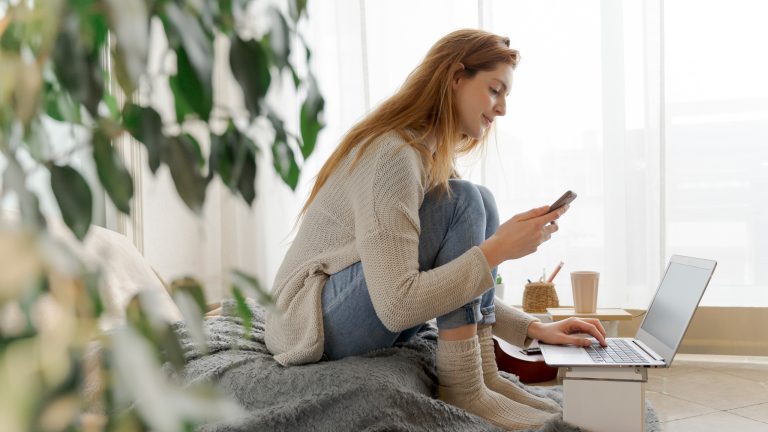 Eine Person sitzt auf einem Bodenkissen und bedient Laptop und Smartphone gleichzeitig.
