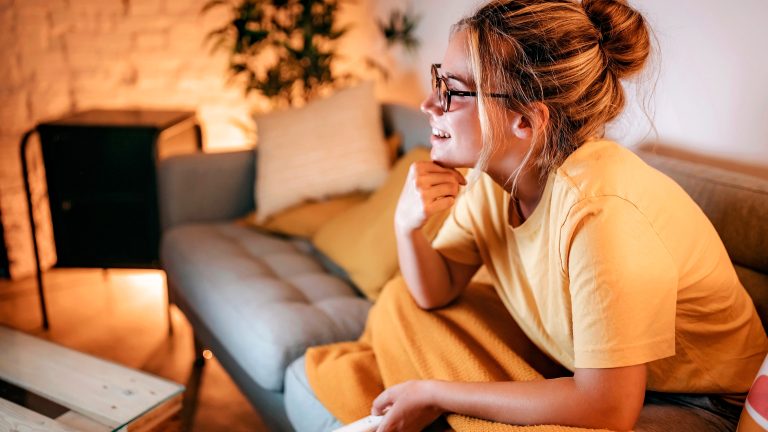Eine Person sitzt auf einem Sofa unter einer Decke und hält eine Fernbedienung in der Hand.