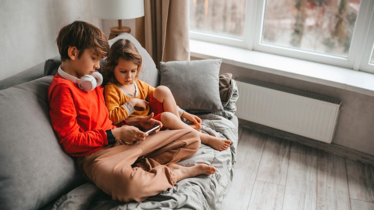 Zwei Kinder sitzen auf einem Sofa und schauen zusammen etwas auf einem Smartphone.