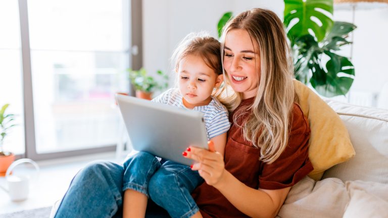 Eine Person sitzt zusammen mit einem Kind auf dem Sofa und schaut in ein Tablet.