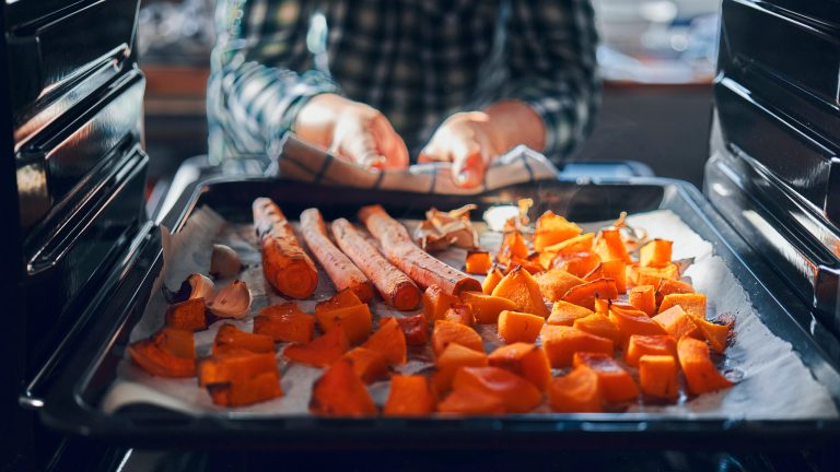 Eine Person nimmt eine Portion Kürbis und Möhren aus dem Backofen.