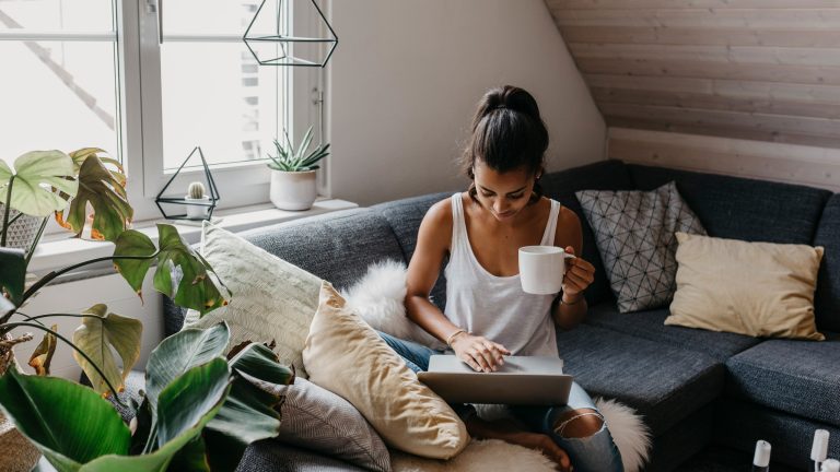 Eine Person mit einem Kaffee in der Hand sitzt auf einem Sofa und bedient einen Laptop.