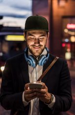 Eine Person mit Kopfhörern um den Hals steht vor öffentlichen Verkehrsmitteln und schaut auf ihr Smartphone.