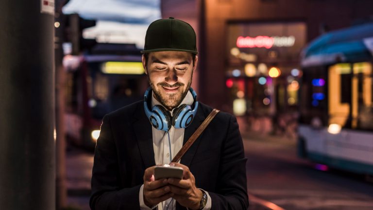 Eine Person mit Kopfhörern um den Hals steht vor öffentlichen Verkehrsmitteln und schaut auf ihr Smartphone.