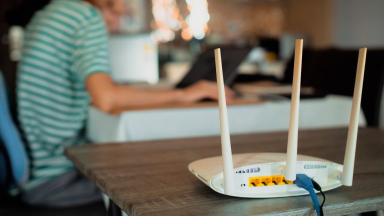 Ein WLAN-Router steht auf einem Tisch. Im Hintergrund ist eine Person an einem Laptop zu sehen.