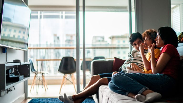 Drei Personen sitzen zusammen auf einem Sofa, essen Popcorn und schauen fern.