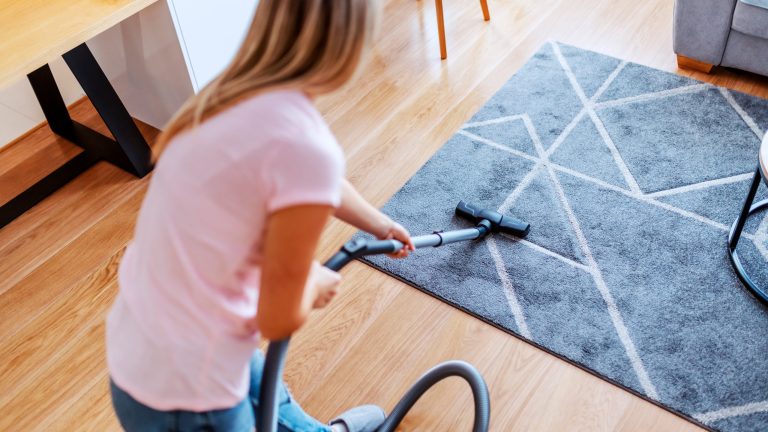 Rear view of attractive worthy caucasian young housewife using vacuum cleaner to clean carpet in living room. Eine Person saugt einen Teppich ab.