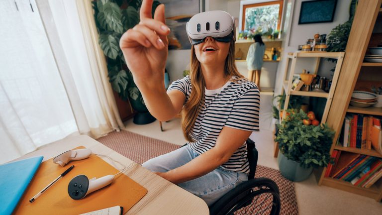 Eine Person sitzt an einem Tisch, auf dem ein Controller der Meta Quest 3 liegt. Die VR-Brille, die die Person trägt, bedient sie mit einer nach oben zeigenden Hand. Eine Person sitzt an einem Tisch, auf dem ein Controller der Meta Quest 3 liegt. Die VR-Brille, die die Person trägt, bedient sie mit einer nach oben zeigenden Hand.