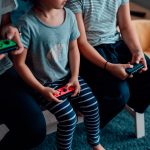 Drei jüngere Personen sitzen auf einem Tisch. In den Händen halten sie jeweils einen Nintendo Joy-Con.