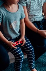 Drei jüngere Personen sitzen auf einem Tisch. In den Händen halten sie jeweils einen Nintendo Joy-Con.